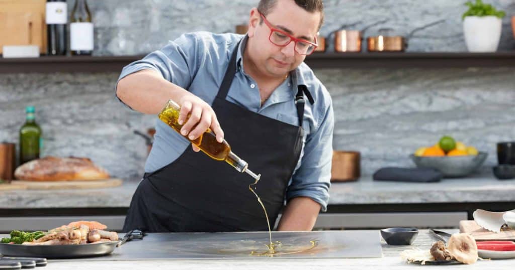 A man wearing an apron pouring olive oil from a glass decanter onto a bbq grill plate.